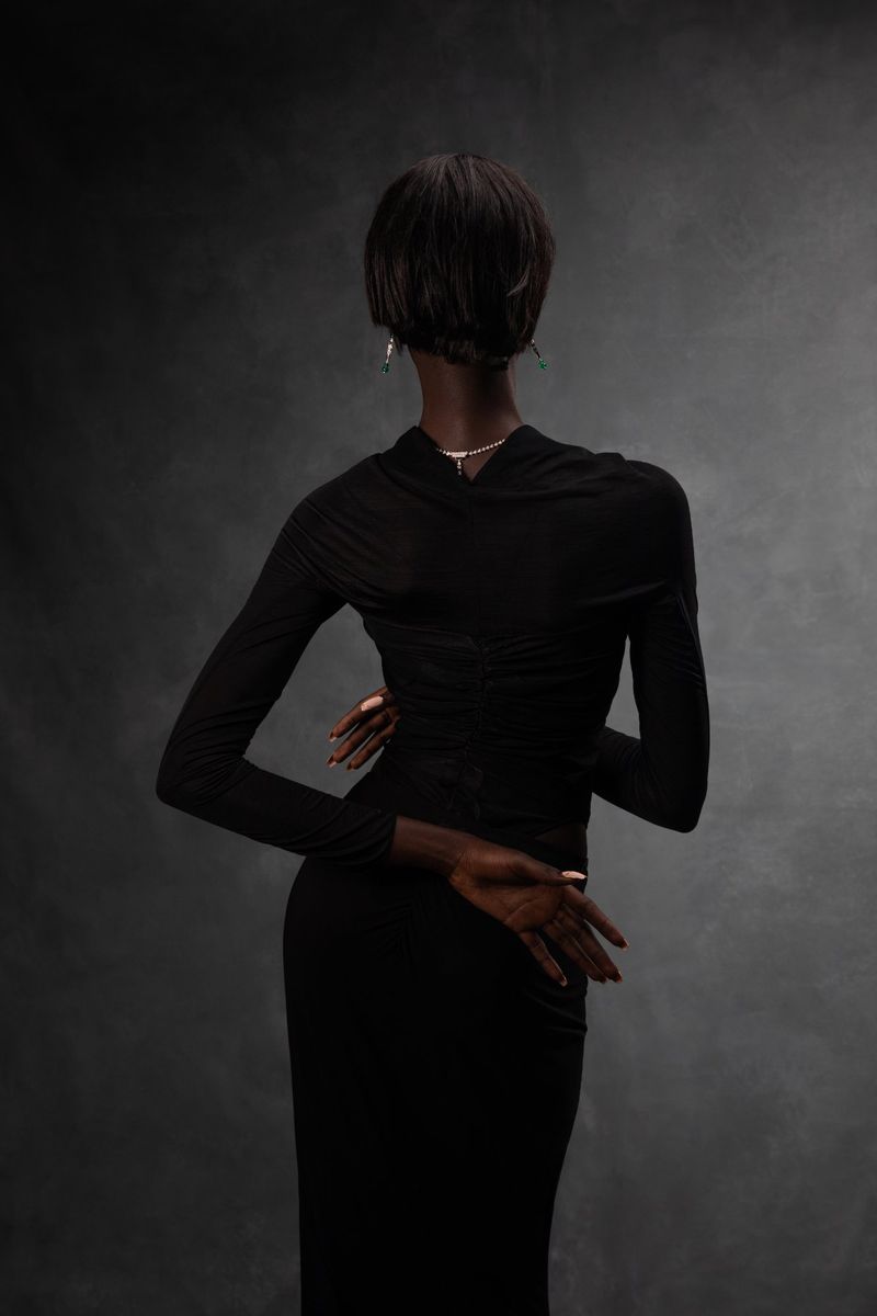 Back view of a slender dark-skinned woman with short black hair wearing a fitted long black dress in a studio setting with grey backdrop.