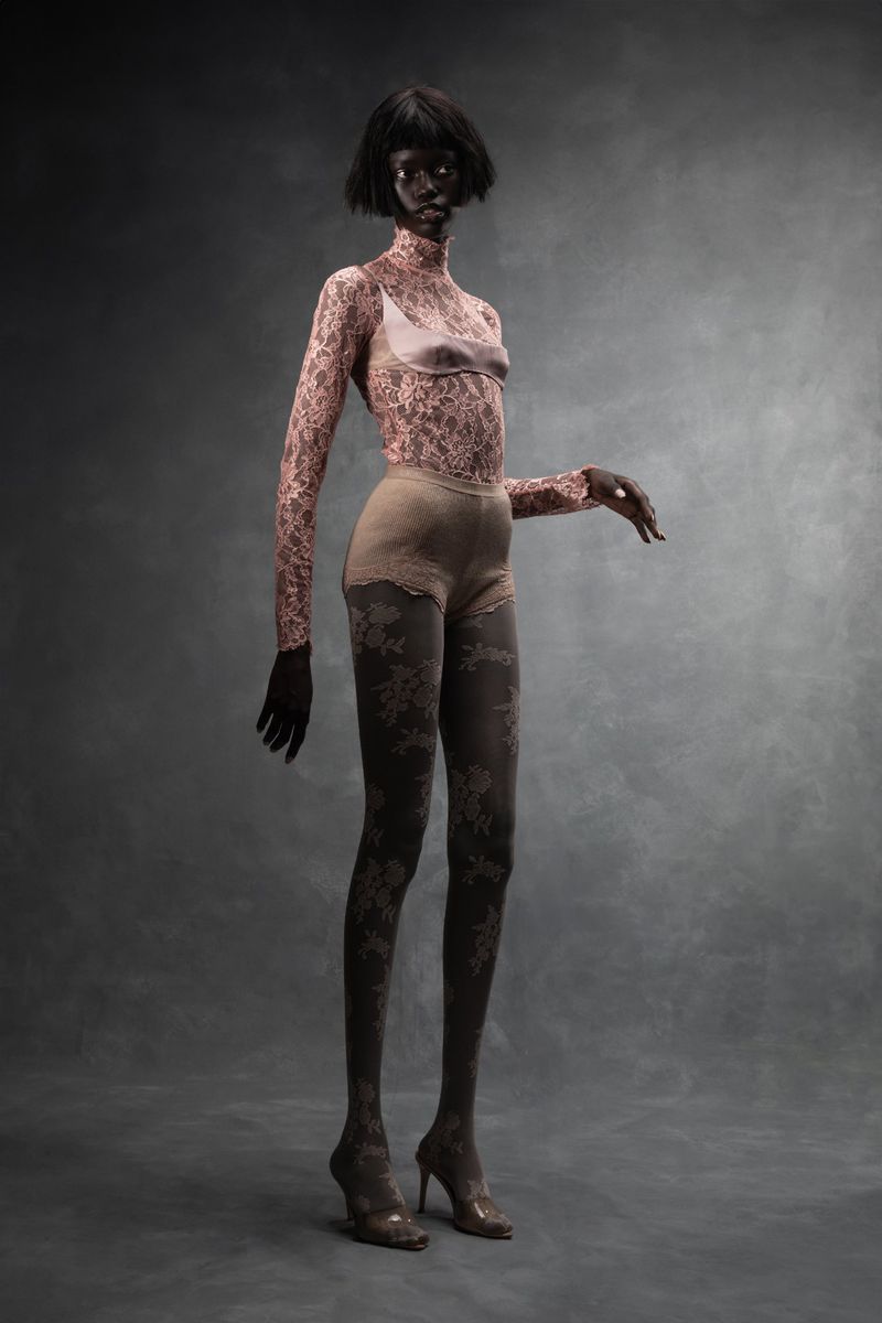A solitary adult woman stands in a three-quarter view wearing a lace top, patterned tights, and heels against a smooth grey background.