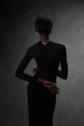 Rear view of a Black woman model in a long-sleeve black dress with hands posed behind her back against a dark grey backdrop.