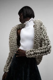 Black model with bob haircut wearing a white ribbed turtleneck and heavy open-knit cardigan against a plain grey studio backdrop.