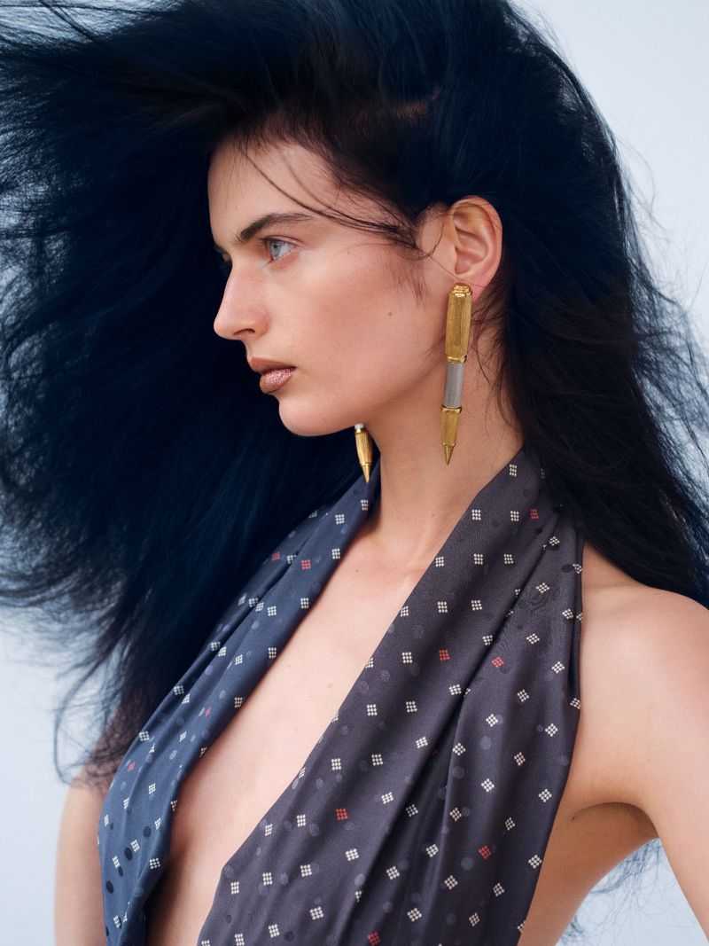 Woman in profile wearing statement earrings and a patterned halter top against a neutral background in a fashion photography style.