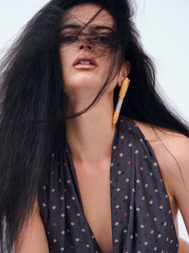 Portrait of a young woman with long dark hair and pen-shaped earrings. She wears a patterned top against a bright background.