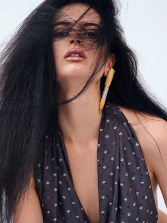 Portrait of a young woman with long dark hair and pen-shaped earrings. She wears a patterned top against a bright background.