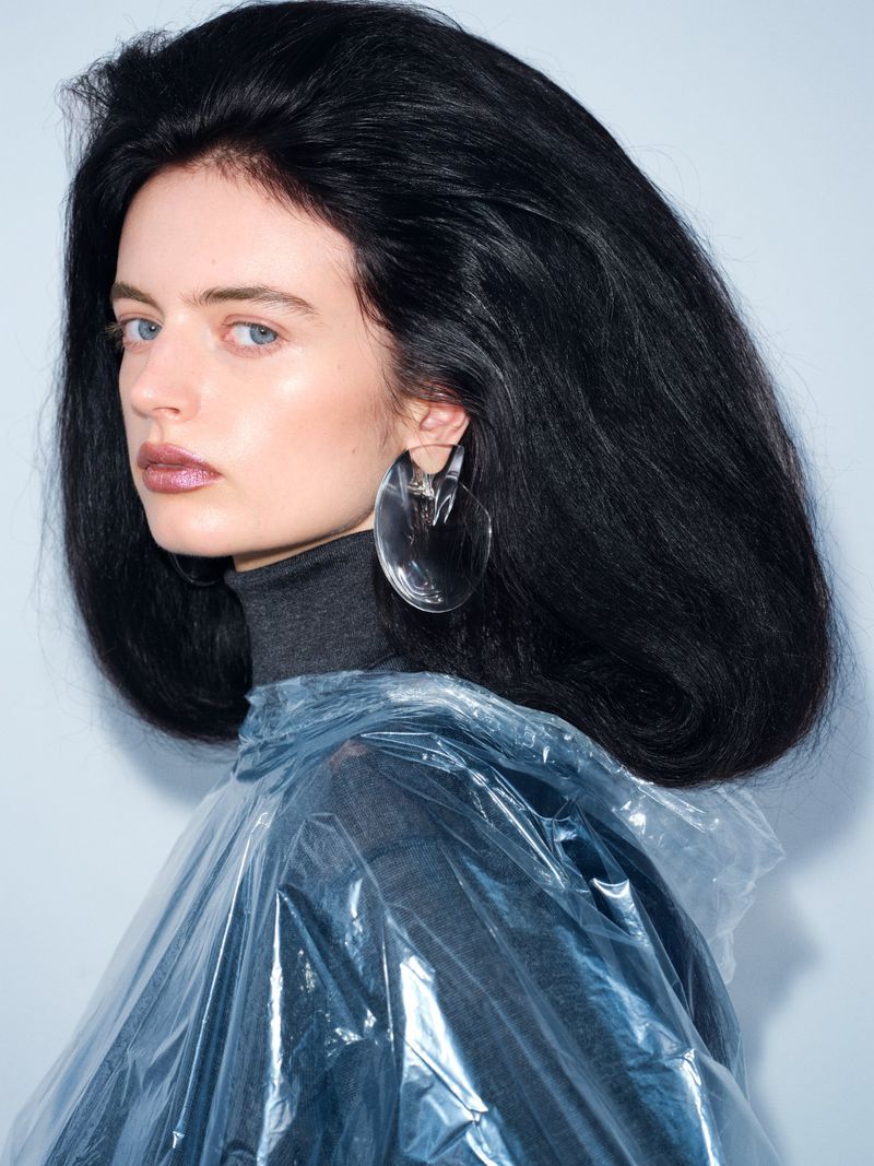 Woman with dark hair and dramatic makeup wearing a gray turtleneck and a clear plastic poncho in a studio setting.