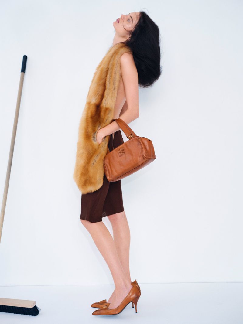 Woman in brown dress and heels posing with a fur stole and leather purse in a clean photo studio, broom in the background.