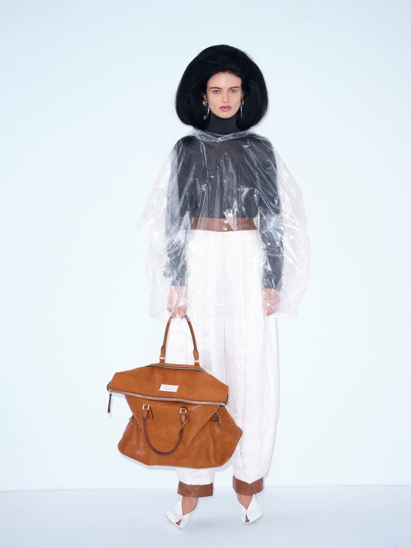Fashionable adult woman poses in photo studio wearing black top, white trousers, clear plastic coat, and brown leather handbag.