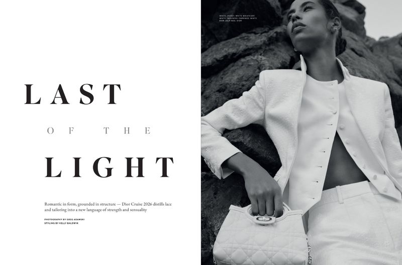 Editorial fashion shot of a Black woman in a white suit posing against a dark rock background, holding a quilted handbag and looking up.