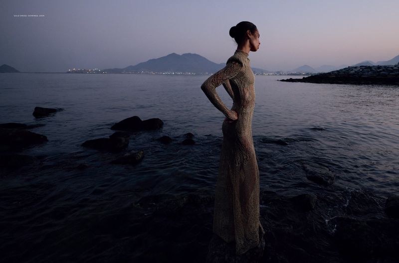 Fashionable woman in a net gown poses on rocks by the water at dusk with mountains and city lights visible in the distance.