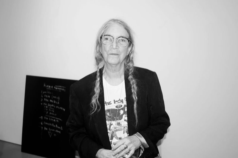 Black and white portrait of Patti Smith wearing glasses, a t-shirt, and a blazer, standing indoors against a white wall.
