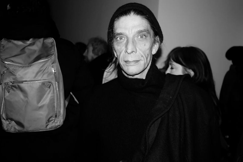 Black and white portrait of an adult male wearing a beanie, surrounded by blurred people, with a backpack visible in the foreground.