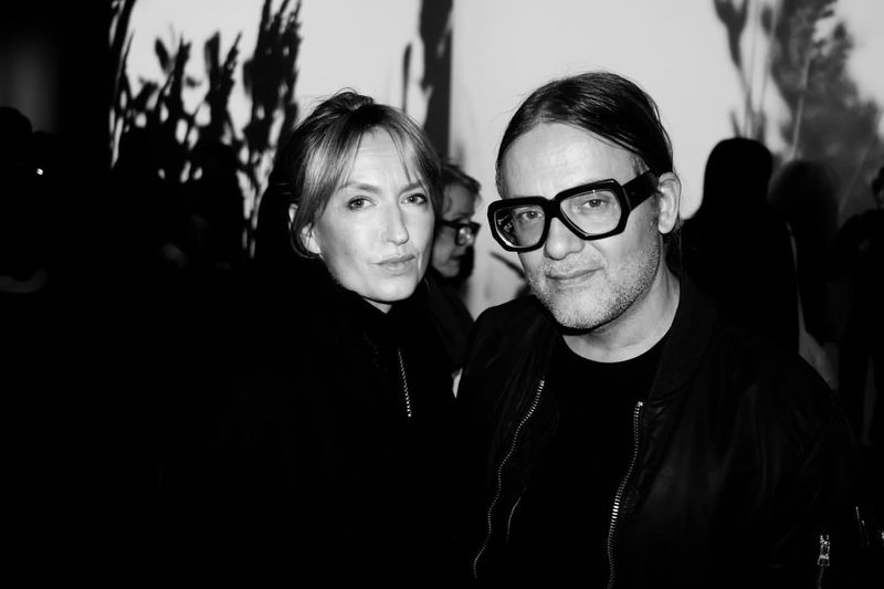 Monochrome portrait of a stylish adult woman and man with bold glasses in front of an abstract projected backdrop, indoor.