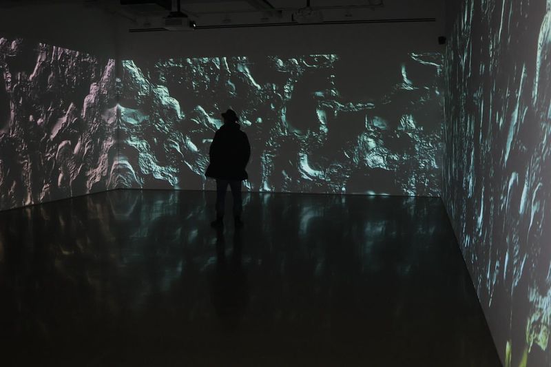A person stands in an art gallery, silhouetted against a wall displaying an abstract projection with textured and patterned effects in light.