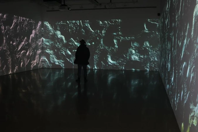 A person stands in an art gallery, silhouetted against a wall displaying an abstract projection with textured and patterned effects in light.