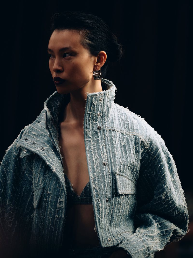 Close-up of an Asian woman in a light blue jacket and matching bra top looking left against a dark backdrop in a fashion setting.