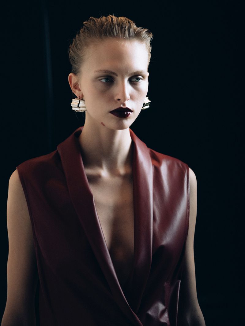 Portrait of an adult woman in a maroon vest with artistic makeup and contemporary earrings against a dark background.