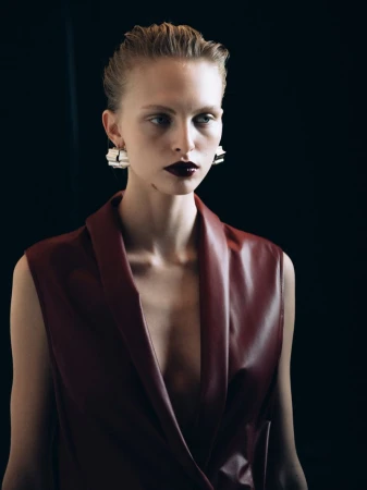 Portrait of an adult woman in a maroon vest with artistic makeup and contemporary earrings against a dark background.