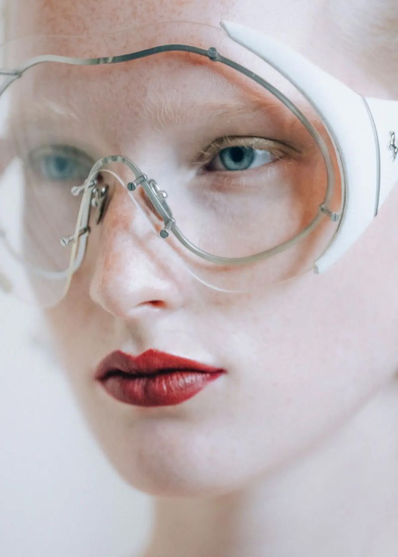 Close-up portrait of a fair-skinned adult female wearing white-framed glasses and dark red lipstick against a bright background.