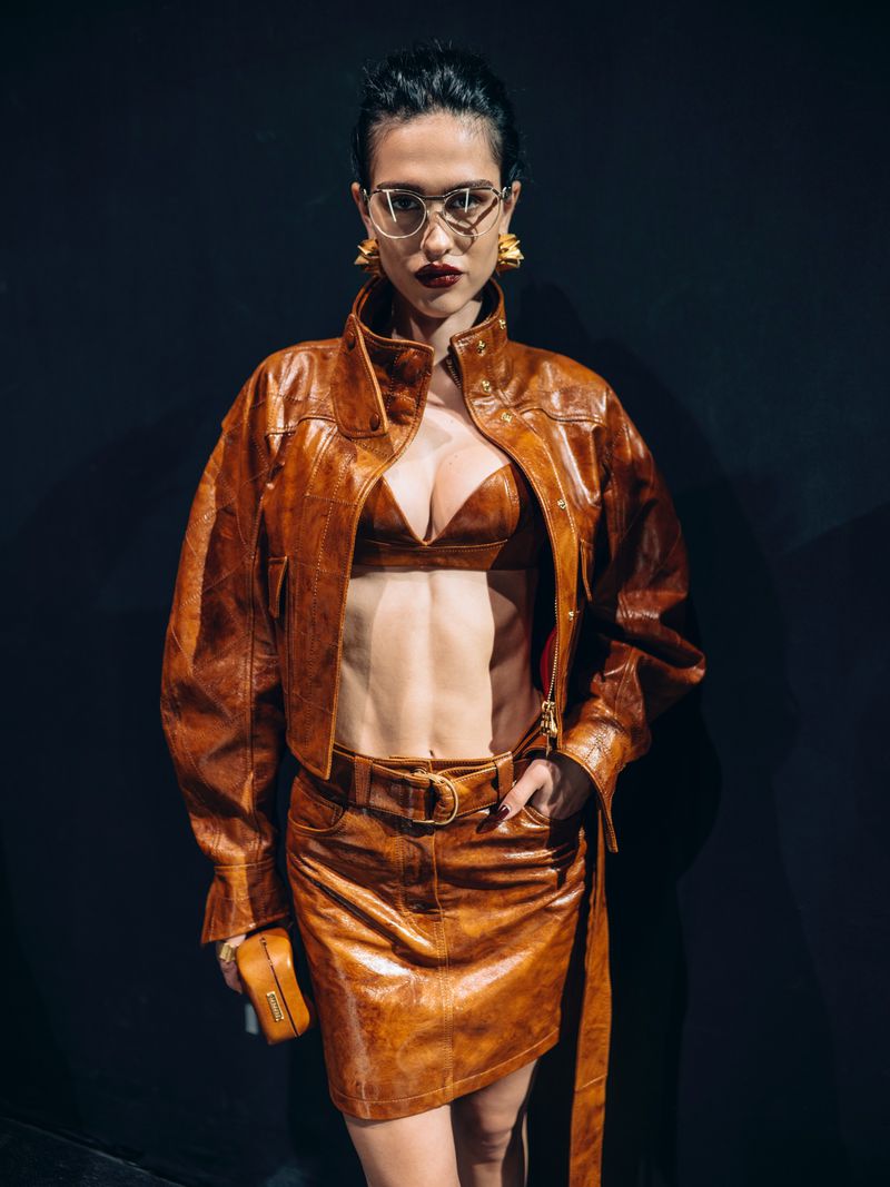 A woman poses in a brown leather bra top, mini skirt, and cropped jacket, completed with gold earrings and glasses against a dark backdrop.