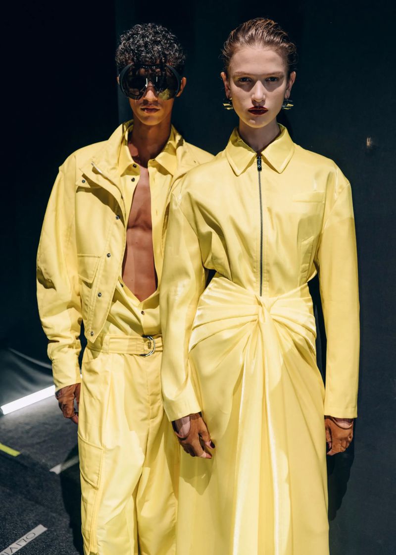 Two fashion models in yellow outfits pose in front of a dark background with hard lighting, highlighting their unique style.