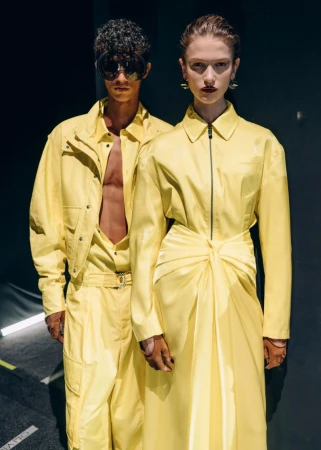 Two fashion models in yellow outfits pose in front of a dark background with hard lighting, highlighting their unique style.