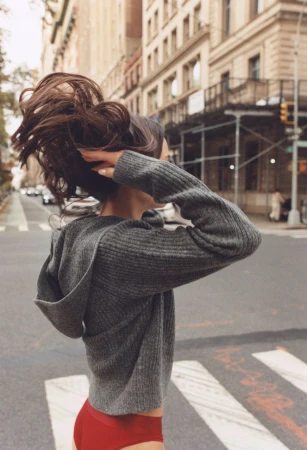 Woman with brunette hair wearing a grey hooded sweater and red bottoms walks across a city crosswalk on a sunny day with buildings in the background.