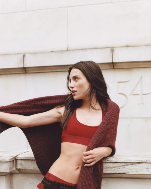 A young woman with long brunette hair poses against a stone wall, wearing a red bra top, cardigan, and underwear, with a Calvin Klein logo.