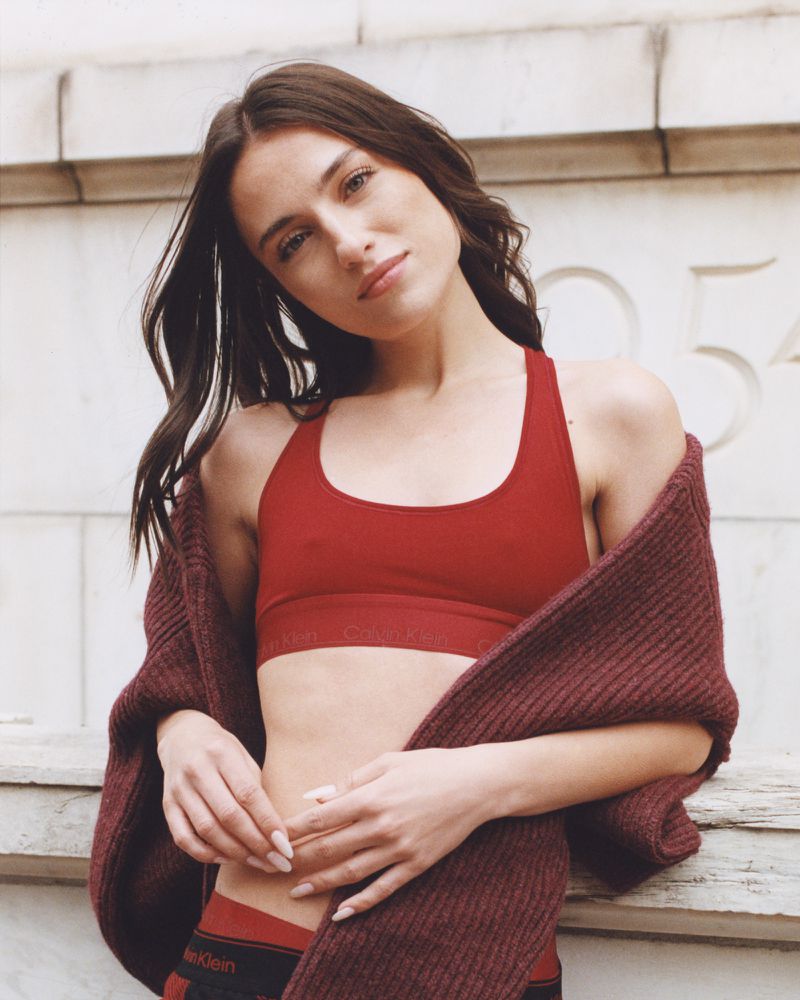 A woman poses in a red sports bra and underwear with a maroon sweater draped on her shoulder, leaning against a weathered wall.