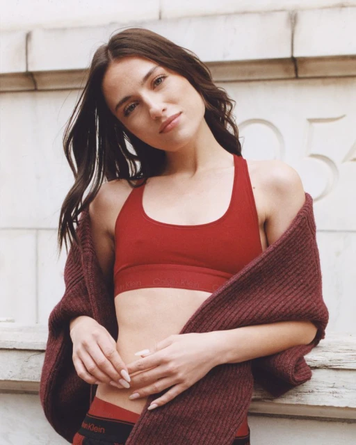 A woman poses in a red sports bra and underwear with a maroon sweater draped on her shoulder, leaning against a weathered wall.