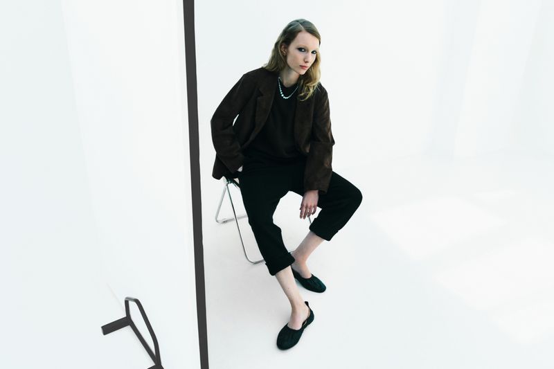 A stylish, tall, slender woman in dark clothing poses while seated on a metal stool in a bright photo studio with a minimalist aesthetic.