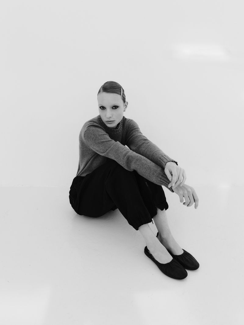 Monochrome fashion shot of a model sitting on a white surface wearing a grey sweater, black pants, and black slip-on shoes.