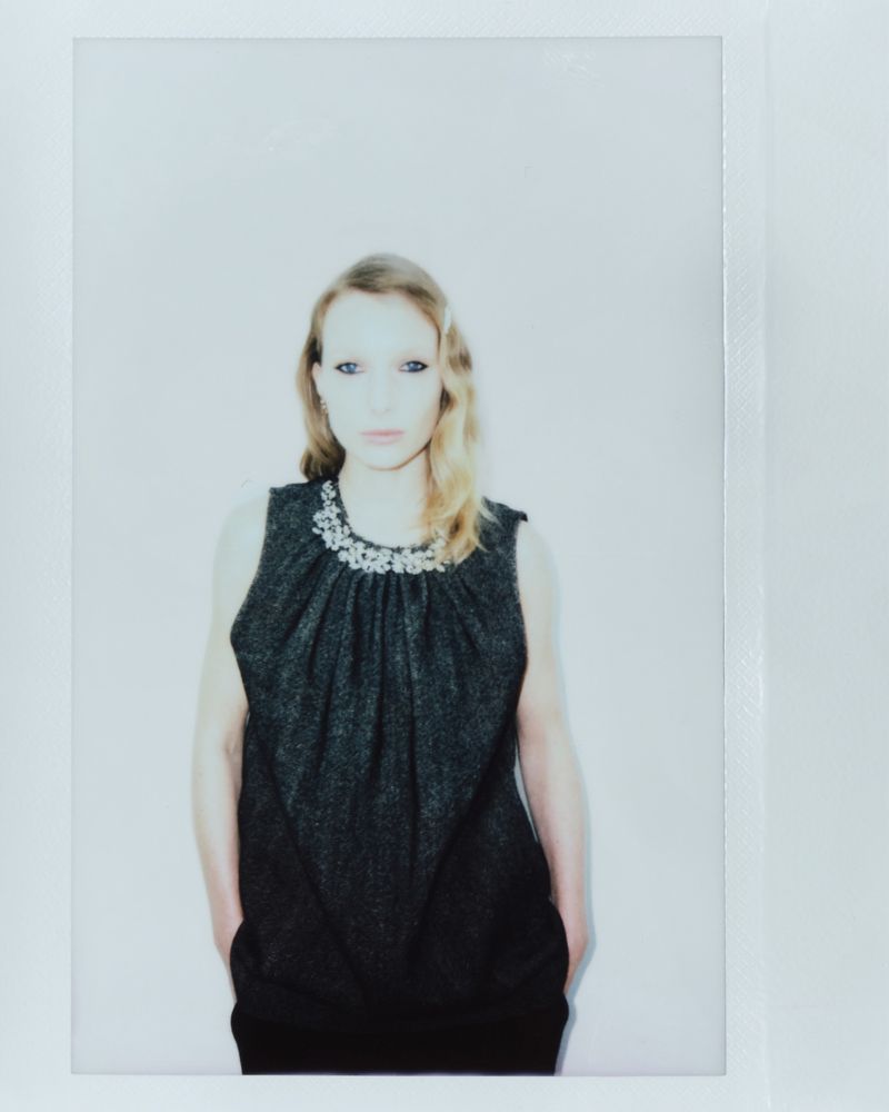 Woman in a grey dress with embellished neckline standing indoors with hands in pockets, captured in a portrait framed by white borders.