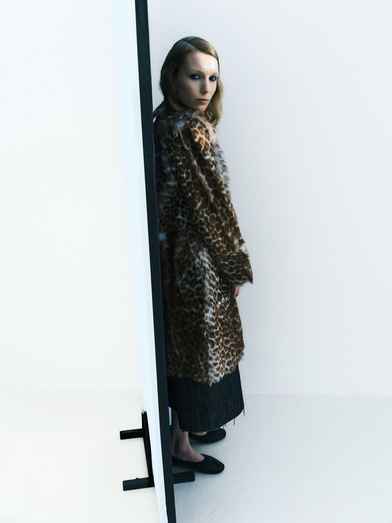 Fashionable woman in a brown leopard print faux fur coat stands in a bright studio by a white panel, posing in black loafers.
