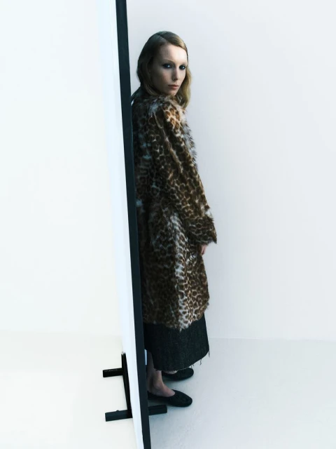 Fashionable woman in a brown leopard print faux fur coat stands in a bright studio by a white panel, posing in black loafers.