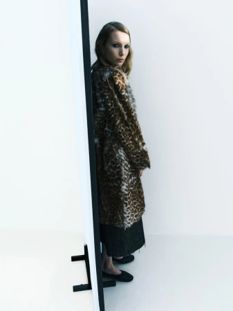 Fashionable woman in a brown leopard print faux fur coat stands in a bright studio by a white panel, posing in black loafers.