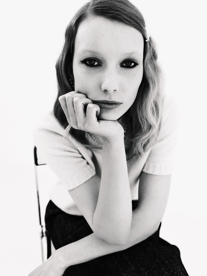 Black-and-white portrait of a young adult female model sitting on a chair, resting her chin on her hand in a photo studio.
