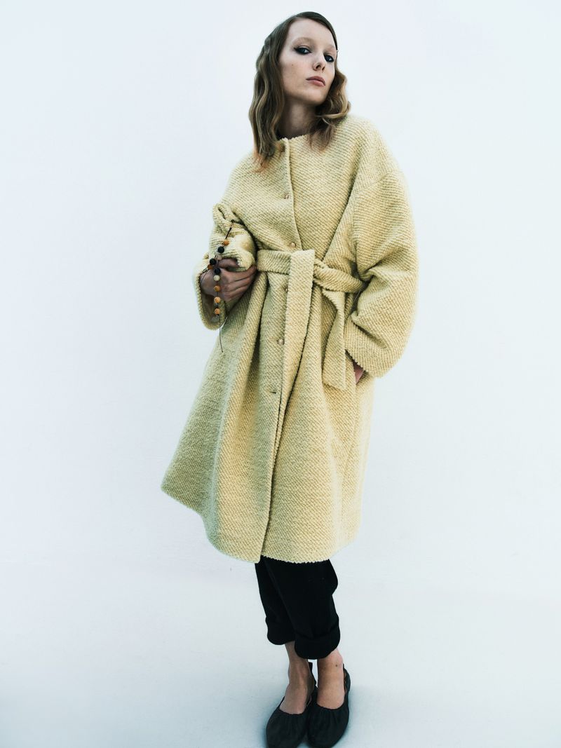 Woman in a textured, belted coat and black trousers, indoors. The model is holding a beaded purse and looking at the camera.