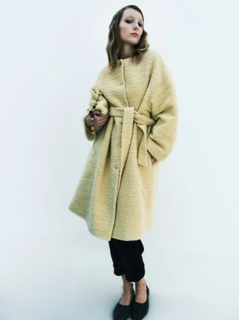 Woman in a textured, belted coat and black trousers, indoors. The model is holding a beaded purse and looking at the camera.