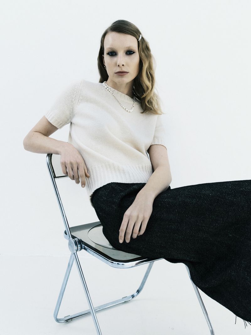 Indoor portrait of a woman with light blonde hair wearing a short-sleeved sweater and black pants sitting on a chrome chair against white background.