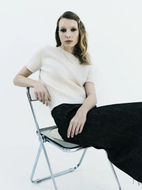 Indoor portrait of a woman with light blonde hair wearing a short-sleeved sweater and black pants sitting on a chrome chair against white background.