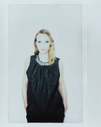 Model wearing a dark sleeveless top with a beaded collar against a plain neutral background in a Polaroid style photograph.