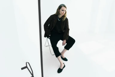 Model seated on a folding chair wearing a dark brown jacket and black trousers against a white seamless backdrop