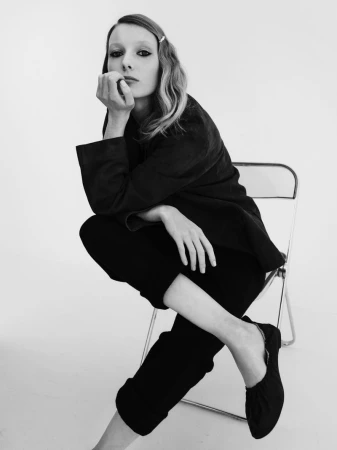 Monochrome portrait of a female model seated on a metal chair against a plain white studio backdrop.