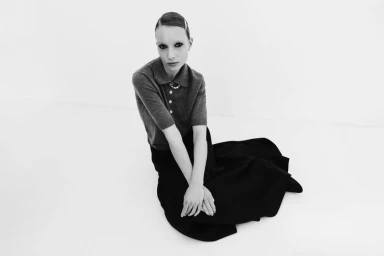 Model seated on white studio floor wearing a dark polo shirt and long black skirt for Rika Studios.