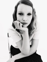 Portrait of a female model resting her chin on her hand, wearing a white top and dark skirt against a white studio background.