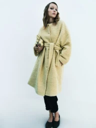 Model wearing a textured yellow belted coat and dark trousers against a white seamless background for Rika Studios.