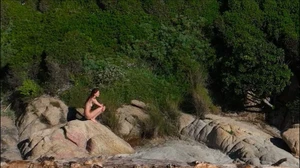 A person crouching on coastal rocky outcrops surrounded by lush green foliage and scrub.