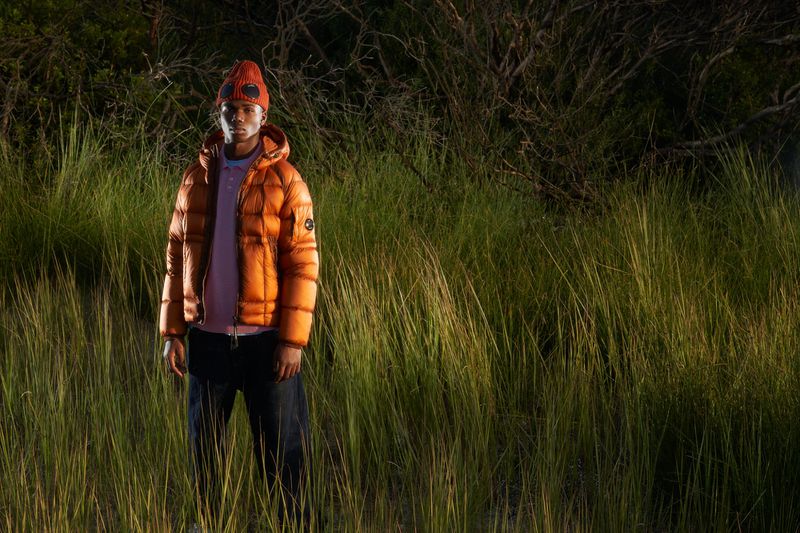 Man in orange puffer jacket and beanie stands in tall grassy field at night, illuminated by artificial light. Fashion and outdoor shot.