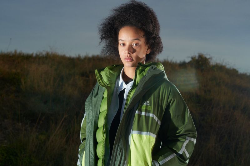 A person with dark curly hair is wearing a green jacket in an outdoor field with a blue sky. The jacket has white stripes. 