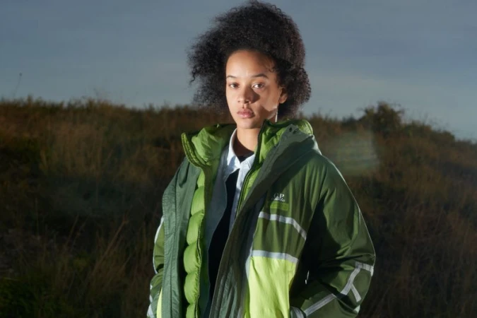 A person with dark curly hair is wearing a green jacket in an outdoor field with a blue sky. The jacket has white stripes. 