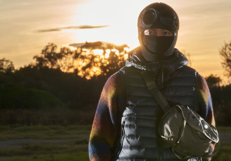 Person wearing black balaclava with goggles, puffer vest, and shoulder bag, standing outdoors against a sunset backdrop.
